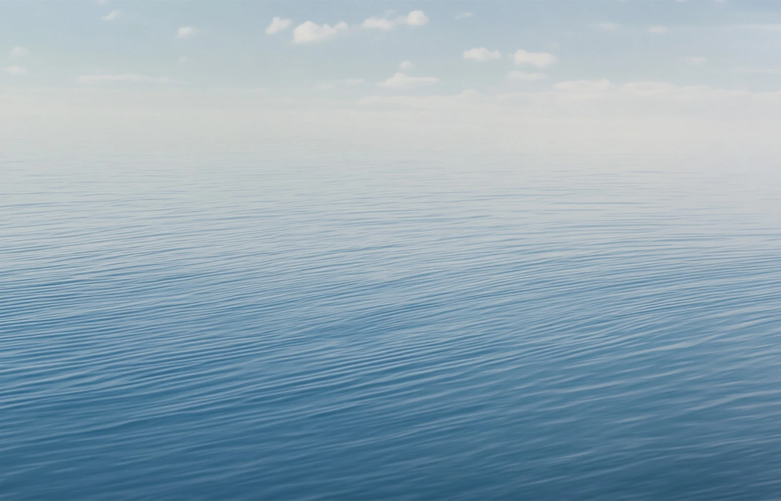 Calm blue ocean with gentle ripples under a pale, cloudy sky and a distant horizon.